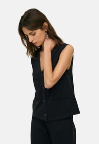 Mujer con cabello castaño hasta los hombros, vistiendo un chaleco negro sin mangas abotonado y pantalones negros a juego, mirando hacia abajo con la mano tocando el cuello.