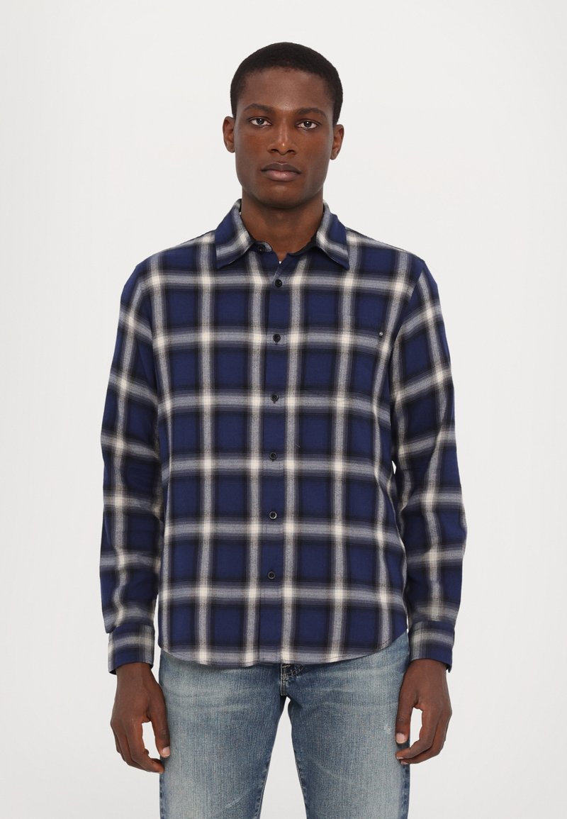 Nöör ja valge ruuduline särk, millel on nööpidega kaelus, pikad varrukad ja veidi kumer hem. Valmistatud tekstuursest kangast.