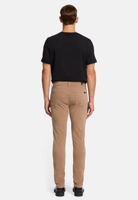 Tan slim fit broek met een zachte textuur, achterzakken en een zwart etiket op de tailleband, gecombineerd met een zwart hemd met korte mouwen.