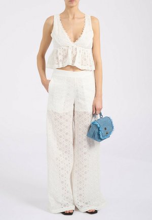 Donna che indossa un crop top bianco in tessuto traforato e pantaloni a gamba larga, tiene una piccola borsa blu testurizzata con dettagli di frange.