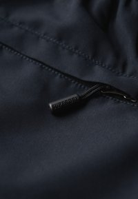 Tela azul marino con una textura suave, presenta un tirador de cremallera negra con "SUPERDRY" grabado. Acentos de costura visibles.