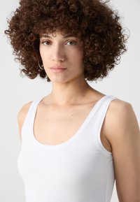 White tank top with a rounded neckline and wide shoulder straps. Smooth cotton fabric. Model has curly, brown hair and is against a light background.
