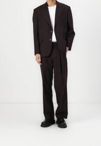 Ruskea räätälöity puku, jossa on muotoiltu jakku, yksittäinen nappisuljenta ja leveälahkeiset housut. Paritettu valkoisen T-paidan ja mustien kenkien kanssa.