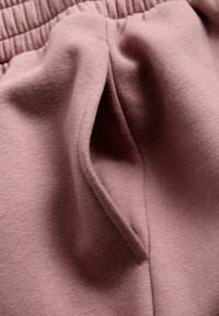 Roze stof met zachte textuur; heeft een taps toelopende vorm en een zijzak. Elastische tailleband zichtbaar aan de bovenkant.