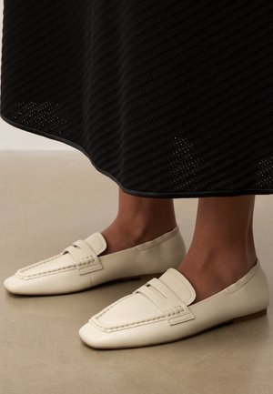 Stopy w kremowych skórzanych mokasynach z przeszyciami, stojące na beżowej podłodze pod czarną fakturowaną spódnicą.