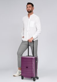 Uomo in camicia bianca e pantaloni grigi in piedi con una mano in tasca e l'altra che tiene il manico di una valigia viola in posizione verticale.