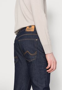Vaqueros de mezclilla azul oscuro con costuras naranjas en contraste, un parche de cuero en la cinturilla y un diseño estándar de bolsillo.