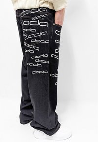 Pantalones de mezclilla negros con un estampado repetido del logo blanco "dada", corte holgado, bolsillos laterales y detalles de costura. Combinados con zapatillas blancas.