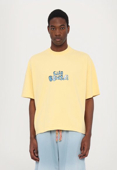Gele oversized katoenen t-shirt met een ronde halslijn en blauwe cartoonachtige tekst "CLUB BONSAI" op de voorkant gedrukt.
