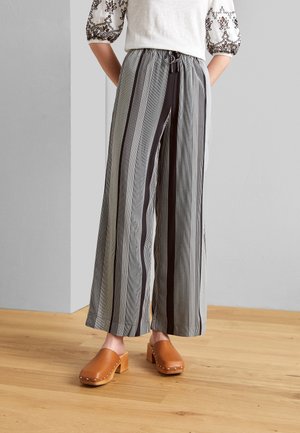 Spodnie o szerokich nogawkach z pionowymi czarnymi, białymi i szarymi paskami, zestawione z brązowymi drewnianymi chodakami na jasnej drewnianej podłodze.