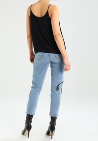 Vrouw in lichtblauwe jeans met zwarte geborduurde patches, een zwarte camisole top en zwarte hooggehakte enkellaarsjes, staand met haar rug naar de kijker gekeerd.