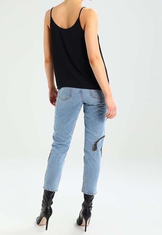 Vrouw in lichtblauwe jeans met zwarte geborduurde patches, een zwarte camisole top en zwarte hooggehakte enkellaarsjes, staand met haar rug naar de kijker gekeerd.