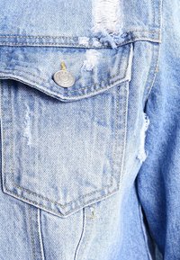 Giacca di denim azzurro chiaro con toppe di usura, dettaglio con bottoni in argento e tasca sul petto. Tessuto testurizzato con cuciture visibili.