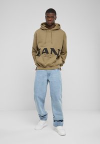 Felpa kaki con grande stampa nera "MAN", cappuccio con coulisse e tasca frontale. Abbinata a jeans baggy azzurri chiari e scarpe bianche.