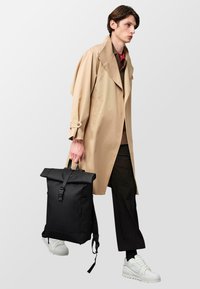 Un sac à dos noir à rabat avec une sangle ajustable, texture lisse, poche zippée sur le côté, porté par une personne en trench-coat beige et baskets blanches.