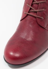 Botín de tobillo de cuero rojo con una textura suave, punta redonda y cierre de cordones en la parte frontal. Cordones marrones suaves y un detalle de diseño minimalista.