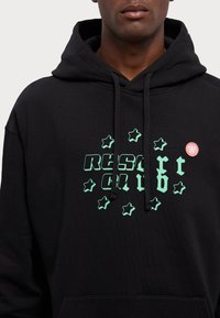 Black hoodie with a front graphic in green featuring the text "RESORT CLUB" surrounded by stars and a red circular logo patch.