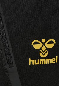 Zwarte sportstof met een verticale rits en een geel logo dat lijkt op een bij met geometrische details. Zachte textuur en slank ontwerp.