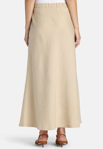 Kvinde iført en beige ankel-lang nederdel med elastik i taljen, kombineret med brune lavhælede sandaler, vist bagfra.