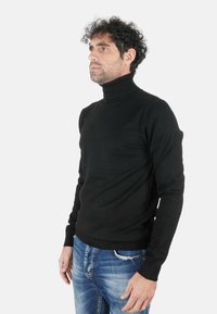 Uomo con capelli ricci che indossa un maglione a collo alto nero e jeans blu, in piedi con il corpo rivolto leggermente verso sinistra su uno sfondo bianco.