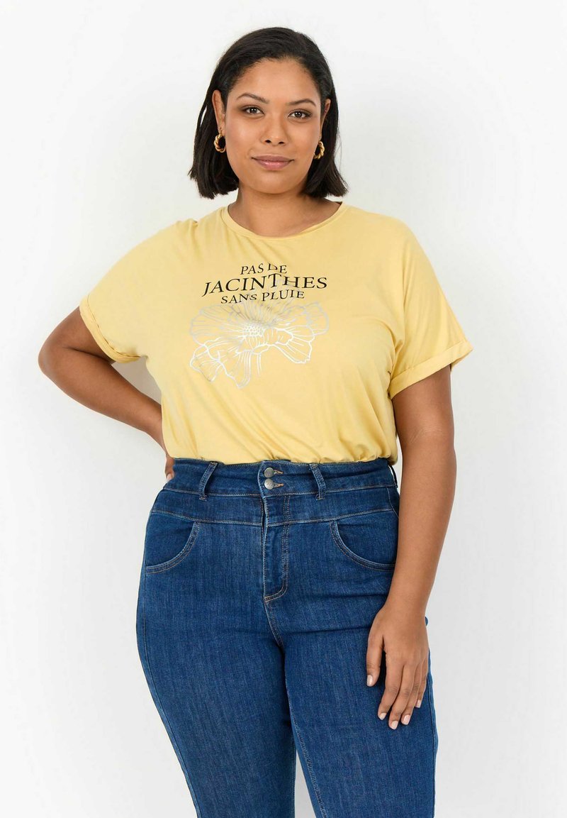 Camiseta amarilla de manga corta con diseño floral y texto. Combinada con jeans de mezclilla azul de cintura alta con dos bolsillos frontales.
