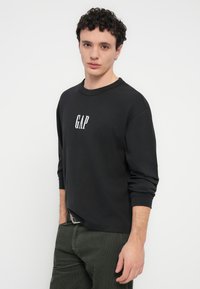 Μαύρο φούτερ με μακριά μανίκια, με λευκό λογότυπο "GAP" στο κέντρο του στήθους. Φορεμένο με παντελόνι από πράσινο σκούρο κοτλέ.