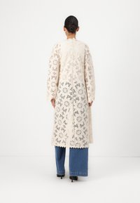 Long, cream lace coat with floral patterns and scalloped edges, worn over blue wide-leg jeans. Back view shows full length and long sleeves.