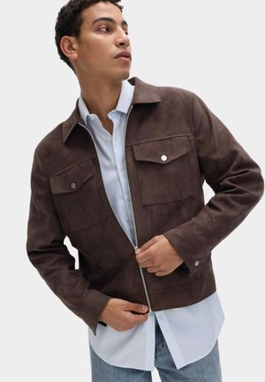 Giovane uomo che chiude la zip di una giacca in camoscio marrone scuro sopra una camicia a righe azzurra e indossa jeans azzurri.