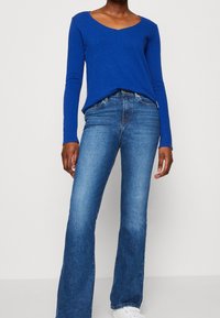 Sininen pitkähihaisen paita V-kauluksella, yhdistettynä loivasti levenevään siniseen farkkuhousuun. Farkuissa on klassinen viiden taskun muotoilu ja vaalea pesu.