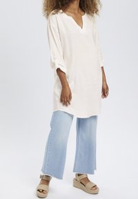 Biała lniana tunika z dekoltem w serek i podwijanymi rękawami, zestawiona z jasnoniebieskimi dzwonami i brązowymi sandałami espadrylami.