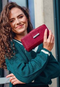 Red leather wallet with embossed texture, a silver clasp, and a zip closure, held against a dark green knitted sweater with white stripes.