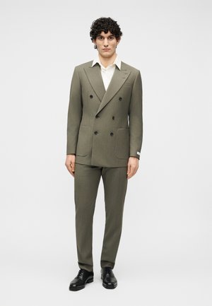 Muž kučeravých vlasov v olivovom dvojradovom obleku, bielom tričku a čiernych spoločenských topánkach stojaci na jednofarebnom svetlom pozadí.