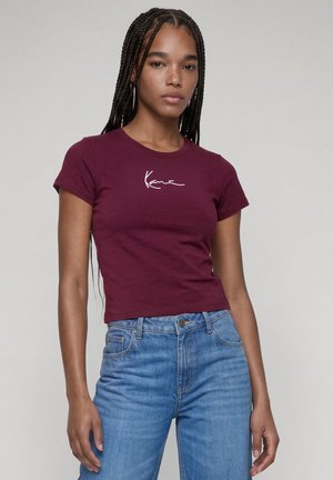 Kvinde med langt flettet hår iført en tætsiddende bordeaux kortærmet skjorte med hvid tekst og højtaljede blå jeans, der står mod en ensfarvet baggrund.