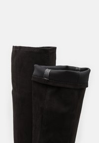 Botas altas hasta la rodilla de color negro, hechas de suave ante, con una parte superior lisa y doblada que presenta una banda de tejido contrastante. Se aprecian detalles de costura.