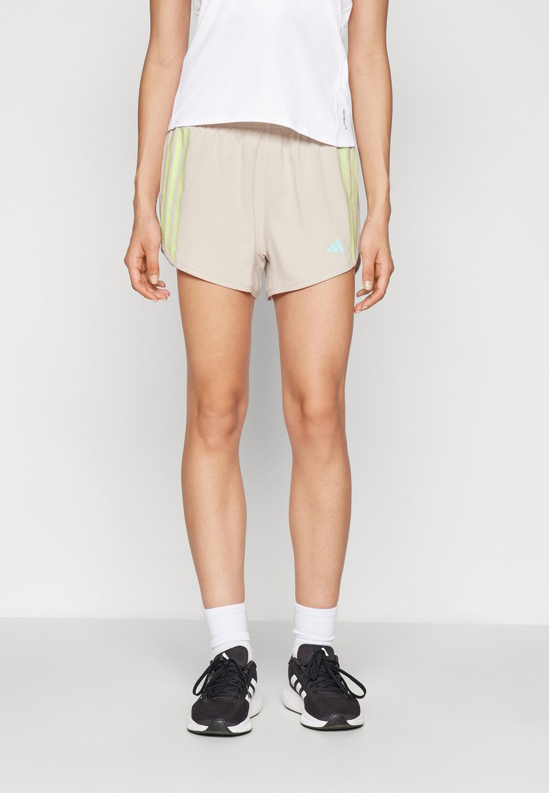 Pantalones cortos deportivos beige con rayas verdes, hechos de un tejido ligero. Presentan un logo de Adidas en azul y un corte relajado, con una longitud por encima de la rodilla.