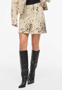 Mini-jupe en denim beige avec motif à pois noirs, présentant un design portefeuille et une fermeture par boutons latéraux. Associée à des bottes stilettos noires montant jusqu'aux genoux.