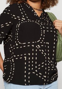 Svart kortärmad topp med v-ringning, prydd med ett krämfärgat vägmönster. Slätt tyg med avslappnad passform. Kombinerad med en grön vadderad jacka.