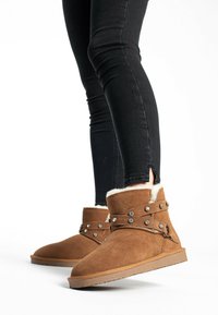 Braune Wildleder-Stiefeletten mit weißem Fellfutter, verziert mit dekorativen Metallnieten und einem Riemen über dem oberen Teil, getragen mit schwarzen Skinny Jeans.