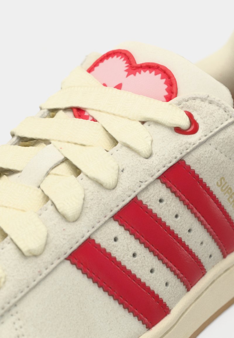 Baskets en daim blanc avec des bandes rouges, des lacets crème et un patch en forme de cœur rose sur la languette. Dotées de détails perforés et d'accents texturés.