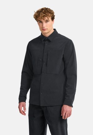 Junger Mann mit lockigem Haar, der ein schwarzes Hemd mit Knöpfen und zwei Brusttaschen sowie schwarze Hose trägt, steht vor einem einfachen hellen Hintergrund.
