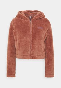 Chaqueta con capucha recortada, con cremallera, de suave y texturizado forro polar de color óxido. Cuenta con capucha y un pequeño parche de logo en el lado izquierdo del pecho.