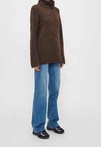 Brauner Strickpullover mit hohem Kragen, kombiniert mit hellblauen geraden Jeans und schwarzen Sneakers mit weißen Akzenten.
