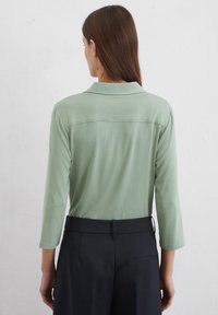 Long-sleeve top in muted green fabric with a collar, paired with dark trousers, showcasing a relaxed fit and simple design lines.