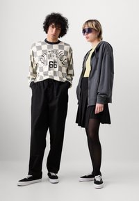 Chemise à manches longues à carreaux avec design graphique, pantalon noir ample, hoodie gris zippé, haut jaune, jupe noire et baskets noires.