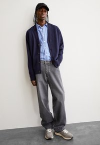 Marine cardigan, met knopen aan de voorkant, gedragen over een blauw gestreept shirt. Losse grijze jeans en grijze sneakers maken de outfit compleet. Eenvoudige pet op het hoofd.