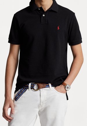 Man in een zwart polo shirt met een rood logo, witte broek, een gestreepte blauw-geel riem, en een bruine leren horloge met een zilveren wijzerplaat.