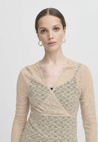 Beige Spitzenoberteil mit langen Ärmeln, Wickeldesign, gewelltem Ausschnitt und Blumenmuster. Verfügt über einen subtilen Glanz und strukturierte Stoffqualität.