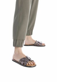 Sandalias tipo slip-on de bronce metálico con un diseño de tira trenzada, suela plana y plantilla suave. Llevadas con pantalones verde oliva con puños.