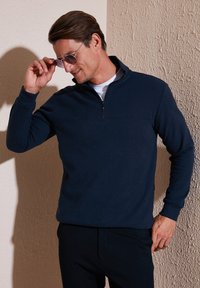 Pullover in pile blu navy con mezza zip e colletto alto. Tessuto liscio con maniche lunghe, indossato sopra una camicia bianca.