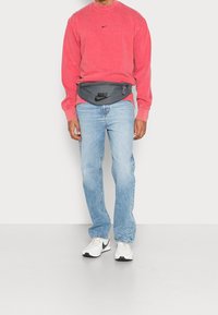 Šedý pumpovací batoh s černým logem Nike, nošený přes červenou mikinu a světle modré džíny, doplněný bílými teniskami s černými akcenty.